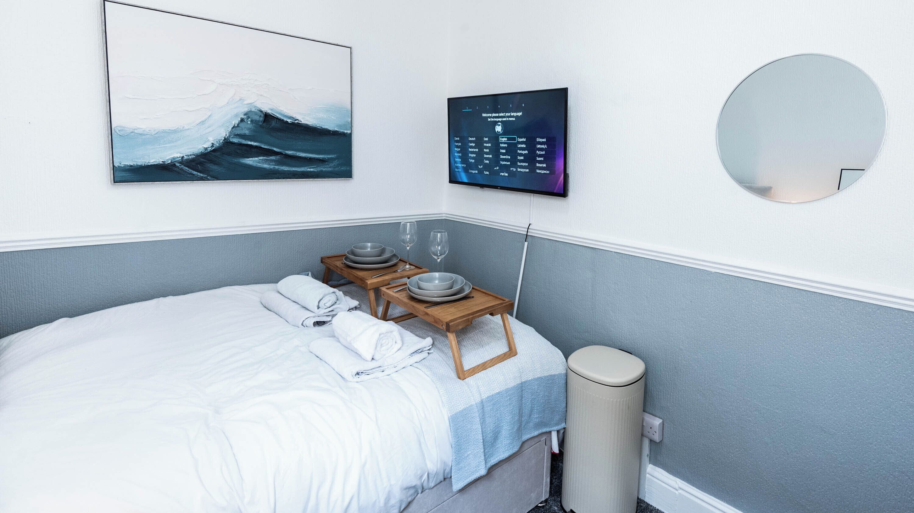 Een hotelkamer met een bed, een nachtkastje met glazen en kommen, een aan de muur bevestigde televisie, een schilderij van een golf en een ronde spiegel.