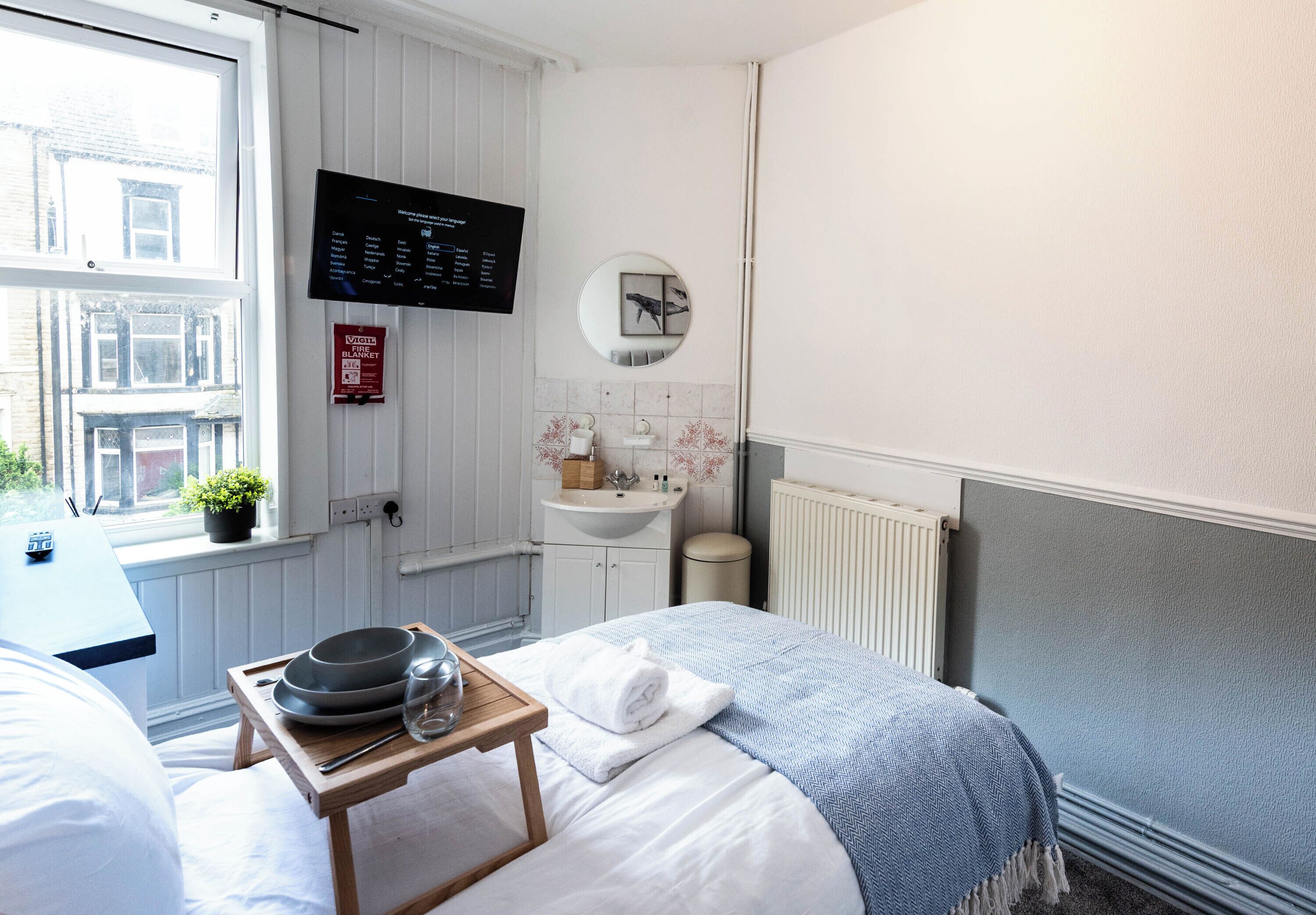 A hotel room with a bed, a bedside table with a tray, a TV, a mirror, a window, a sink, and a radiator.