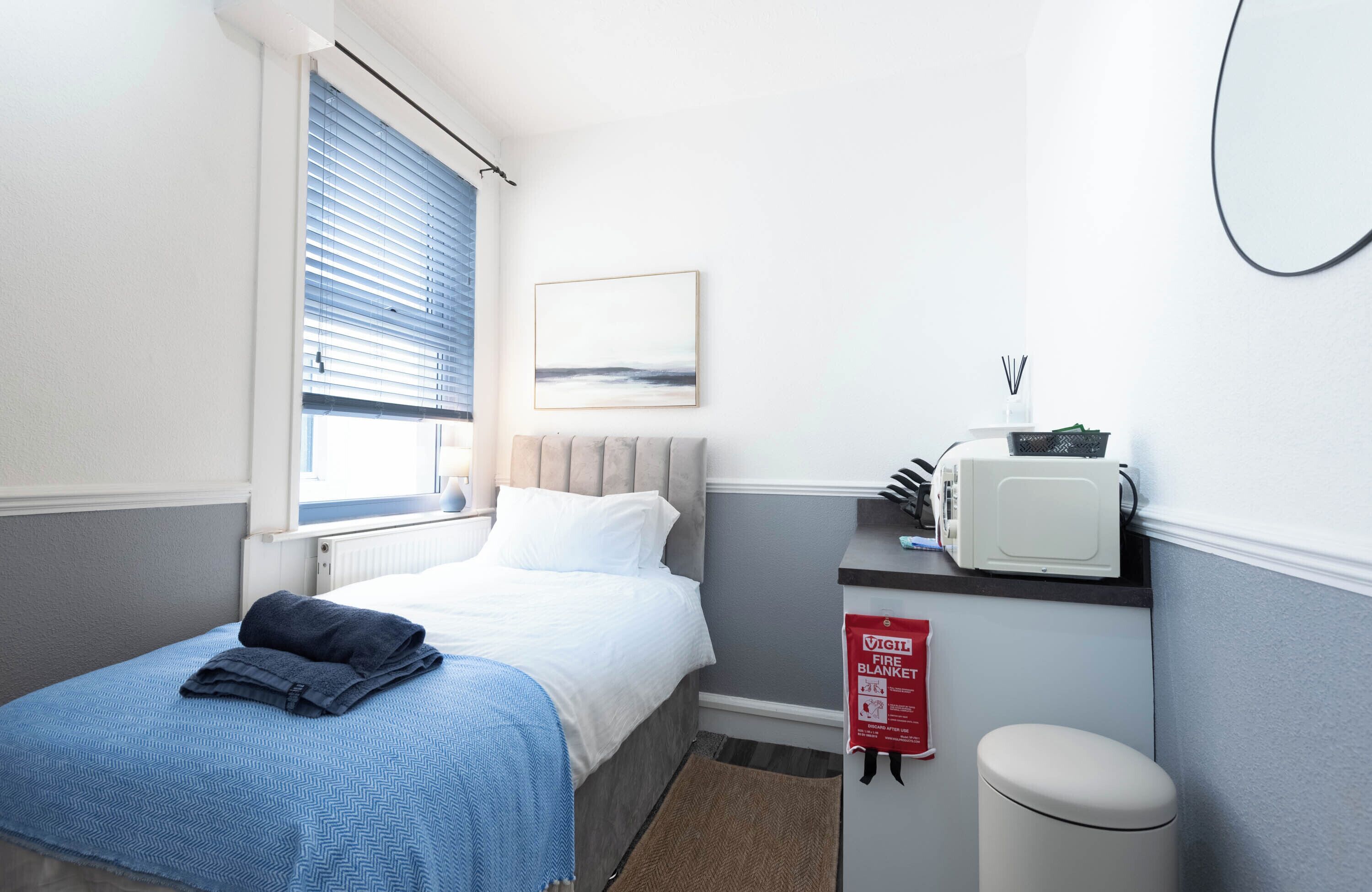 A hotel room with a bed, a window with blinds, a wall-mounted fire blanket, a white trash bin, and a small table with a microwave.