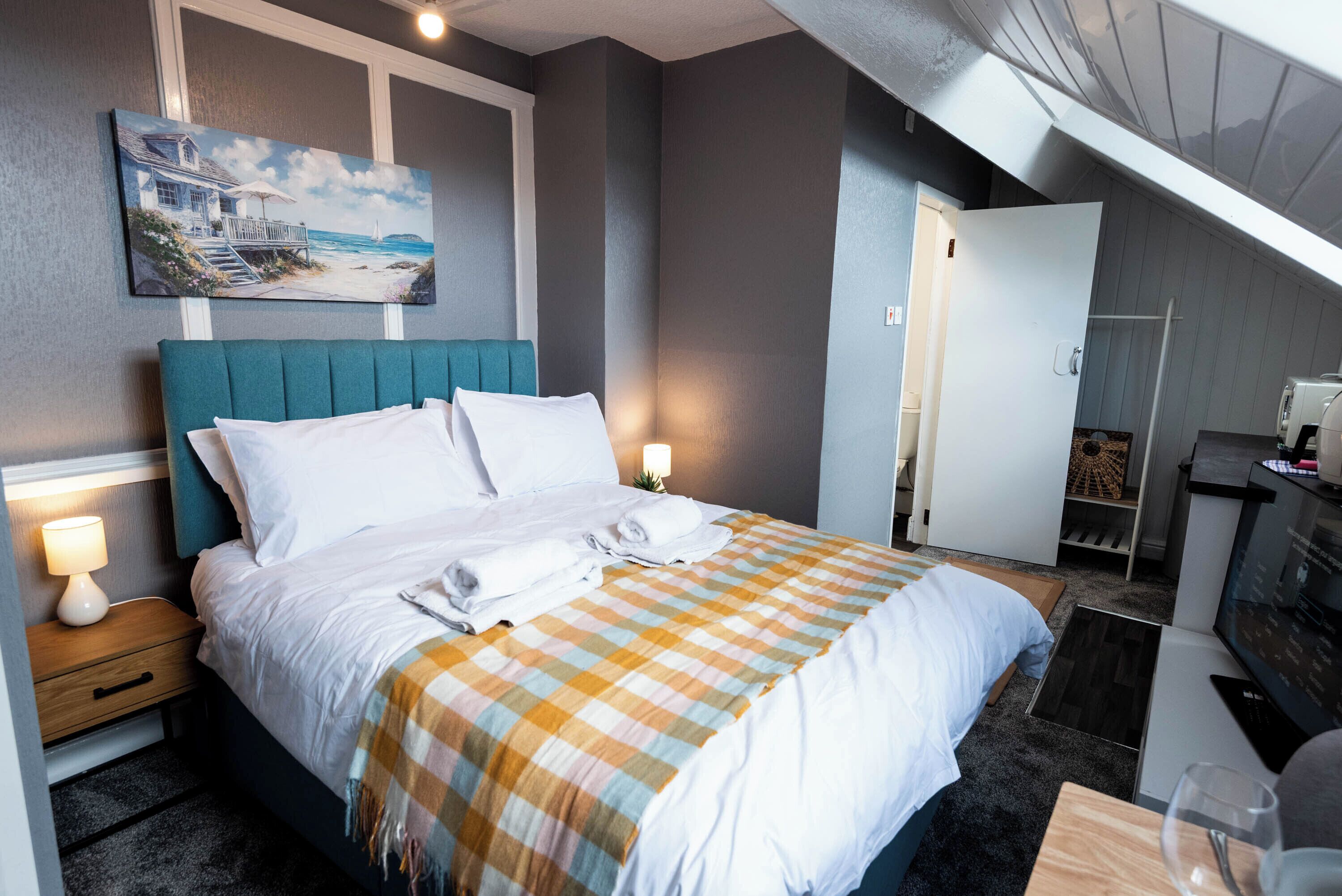 A hotel room with a bed, a painting on the wall, a desk with a computer, and a door leading to another room.