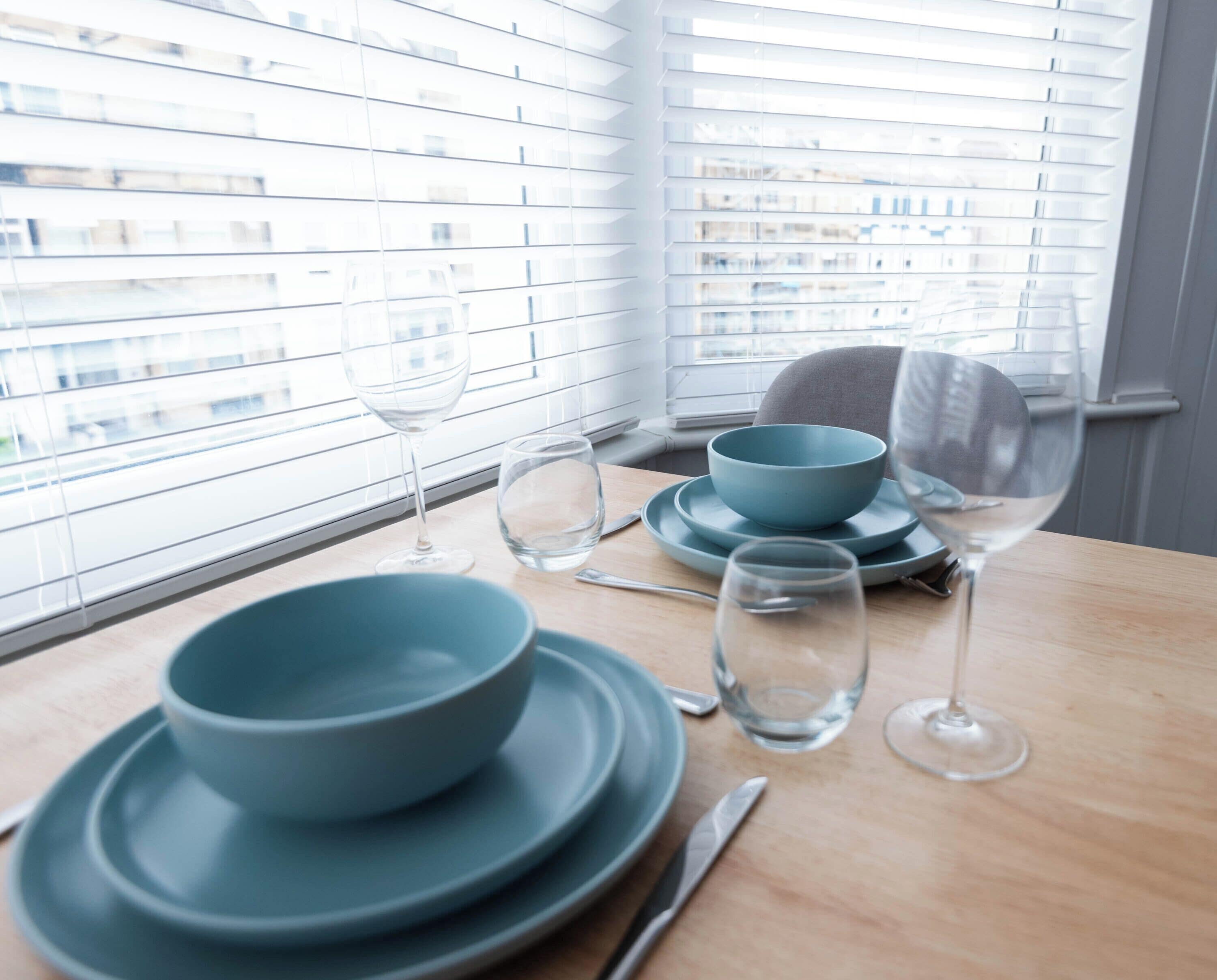 A dining table with blue plates, a bowl, and wine glasses, set against a window with blinds.