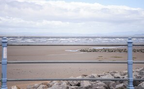 A beach with a metal railing in the foreground, a rocky shoreline, and waves in the background. - The Bay by Guestz (Morecambe)