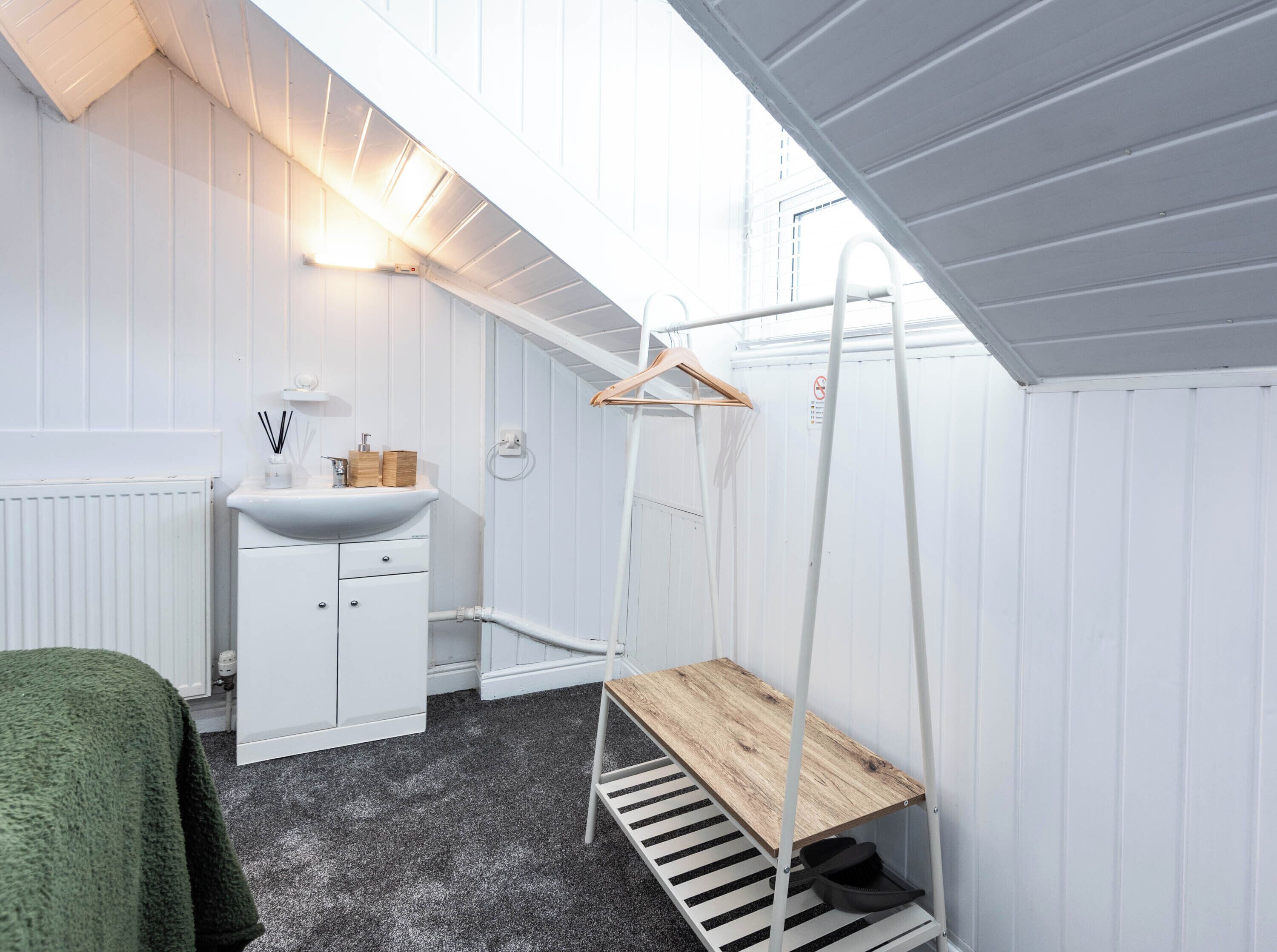 Una habitación pequeña con paredes blancas, un radiador, un lavamanos, una estantería de madera y un perchero.