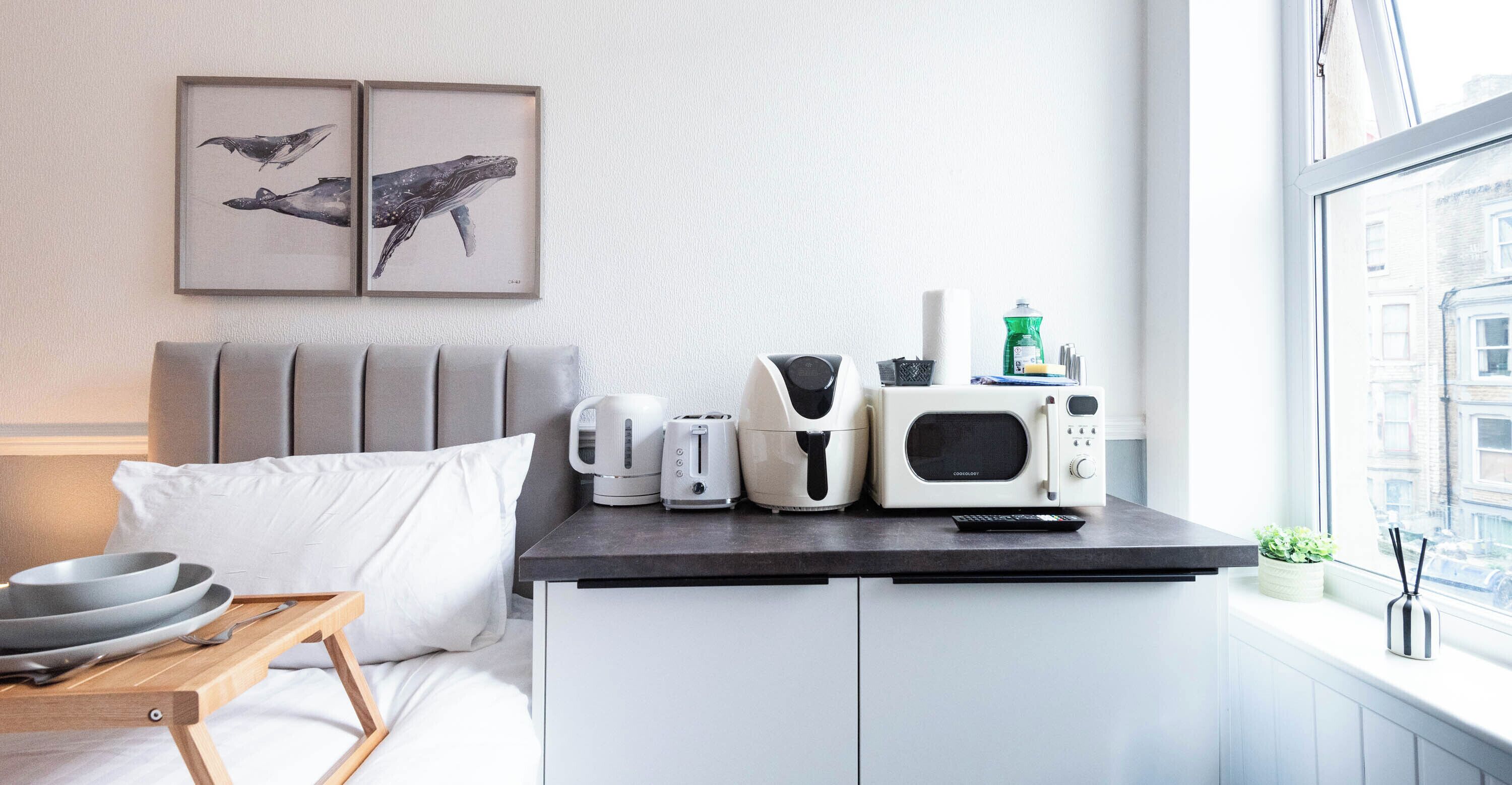A modern hotel room with a bed, a small kitchenette with a microwave, toaster, kettle, and a coffee maker. There are two framed whale paintings on the wall, and a window with a view of buildings.