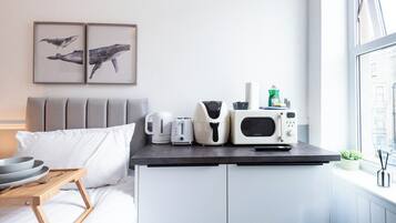 A modern hotel room with a bed, a small kitchenette with a microwave, toaster, kettle, and a coffee maker. There are two framed whale paintings on the wall, and a window with a view of buildings.