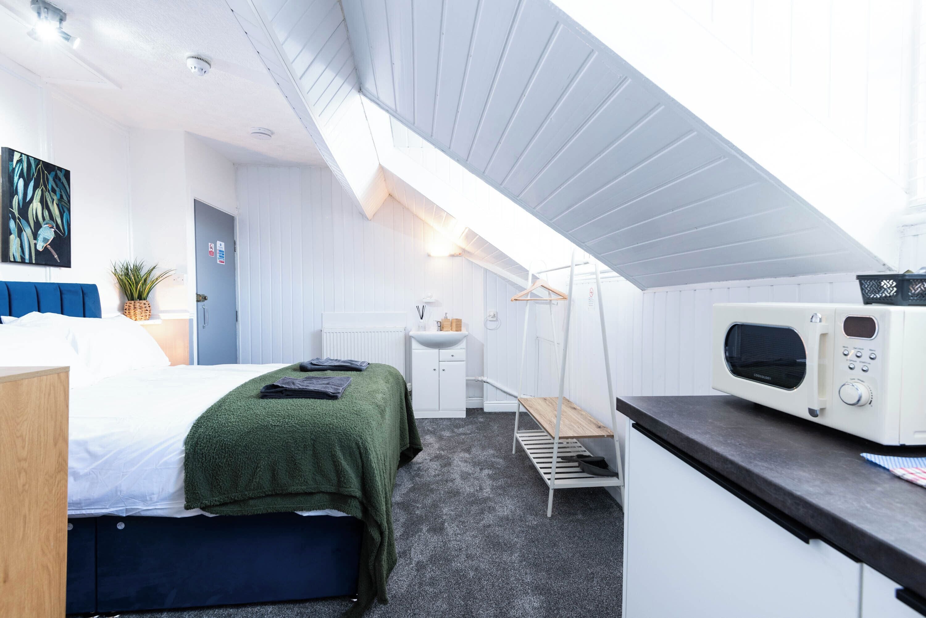 A compact hotel room with a bed, a small kitchenette, a hanging chair, and a ceiling fan.