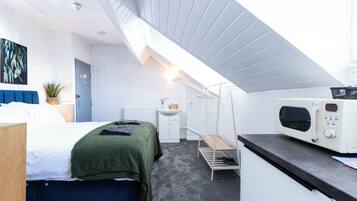 A compact hotel room with a bed, a small kitchenette, a hanging chair, and a ceiling fan.
