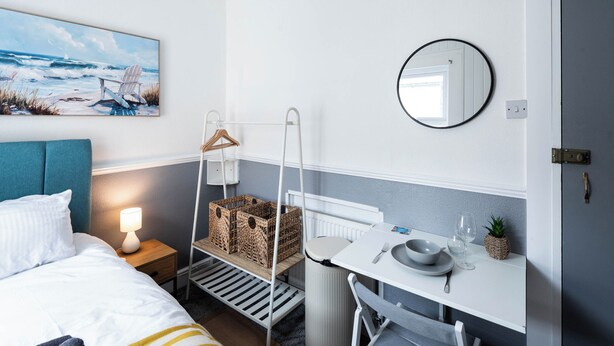 Ein Hotelzimmer mit einem Bett, einem Nachttisch mit Lampe, einer hängenden Pflanzkorb, einem kleinen Tisch mit Schüssel und Glas, einem Spiegel und einem Bild von einer Strandlandschaft.