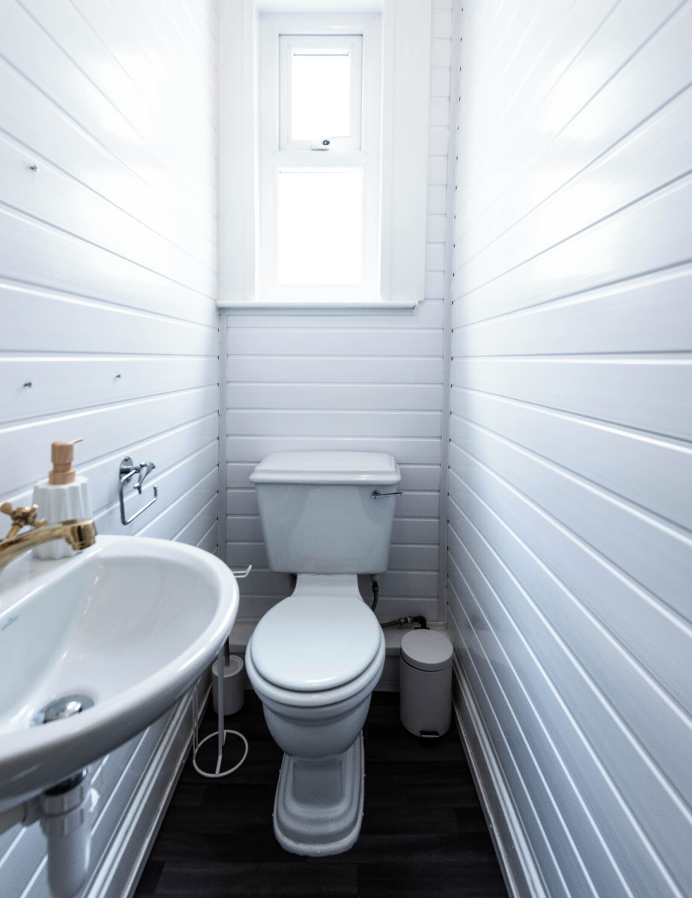 Un baño pequeño con inodoro blanco, lavamanos con grifo dorado y ventana sobre el inodoro.