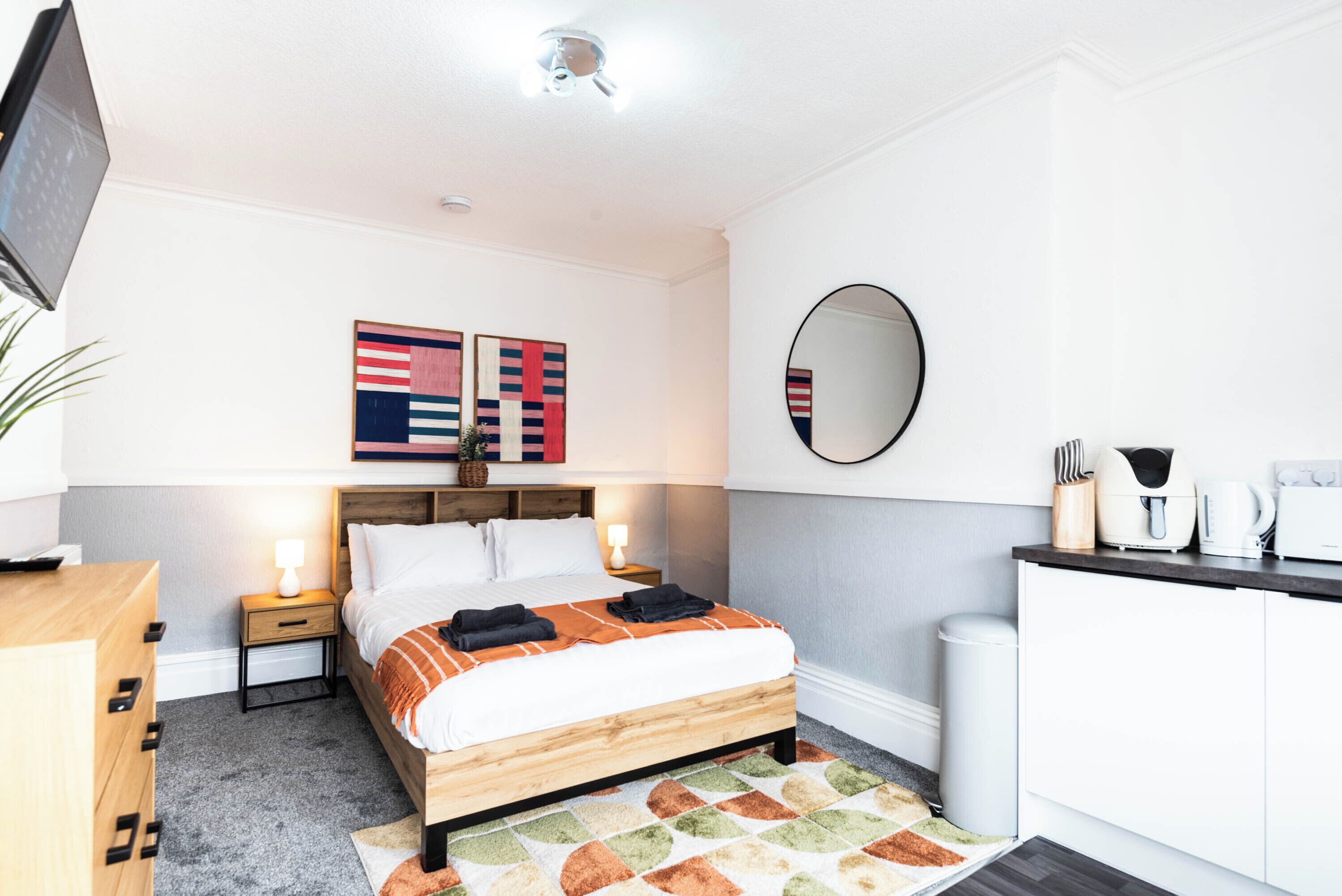 Un dormitorio moderno con cama, mesitas de noche, televisor, espejo y kitchenette.