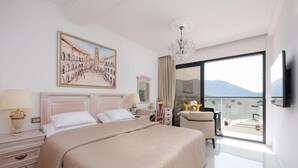 Ein Hotelzimmer mit einem großen Bett, einem Stadtbildgemälde, einer Kristallleuchtre, einem Balkon mit Blick und einem Flachbildfernseher.
