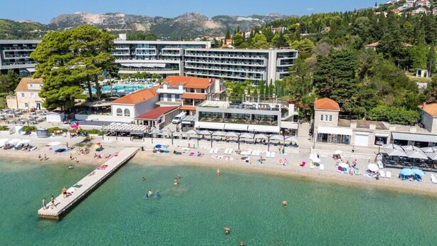 Ein Hotel an der Küste mit mehreren Etagen, einer in das Meer hineinragenden Mole und einem Strandbereich mit Sonnenschirmen und Liegestühlen.