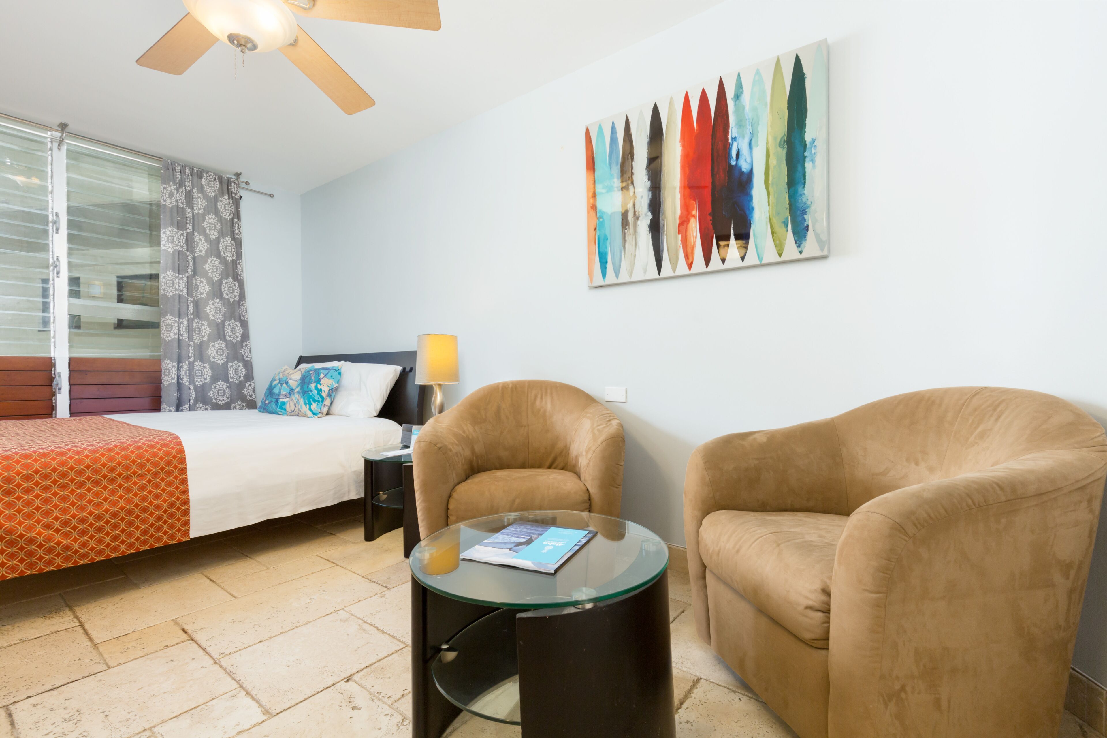 A hotel room with a bed, two armchairs, a glass coffee table, a lamp, and a painting on the wall.