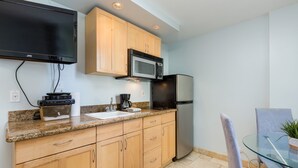 A kitchen with wooden cabinets, a microwave, a refrigerator, a glass table, and a television mounted above the counter. - Studio w/Kitchenette, AC, WiFi, & Flat Screen - Waikīkī Grand #400 (Honolulu)