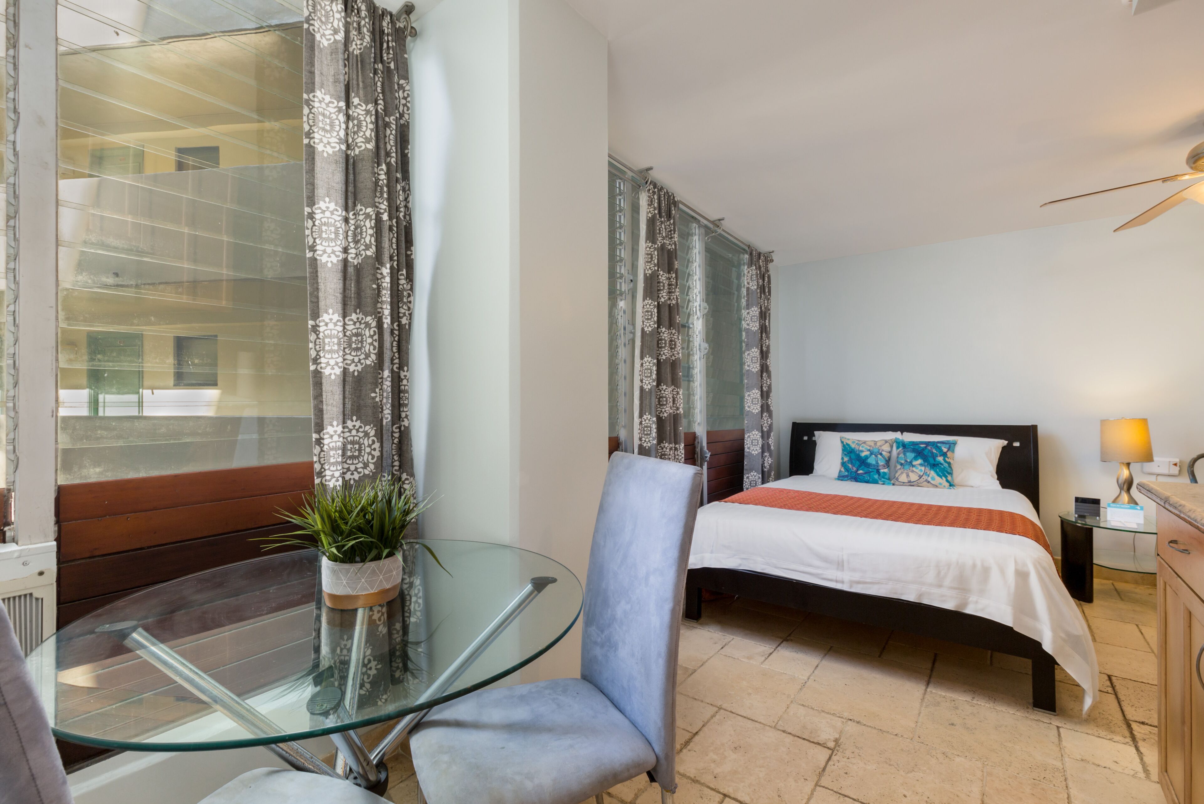 Ein Hotelzimmer mit Glastisch, einem Sessel, einem Bett mit weißer und orangefarbener Bettdecke und Vorhängen mit Blumenmuster.
