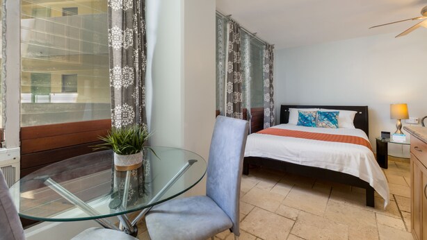 A hotel room with a glass table, a chair, a bed with a white and orange bedspread, and curtains with a floral pattern. - Studio w/Kitchenette, AC, WiFi, & Flat Screen - Waikīkī Grand #400 (Honolulu)
