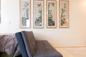 Ein Zimmer mit grauem Sofa, einem Bett mit gemusterten Betttextilien und drei gerahmten Kunstwerken an der Wand.