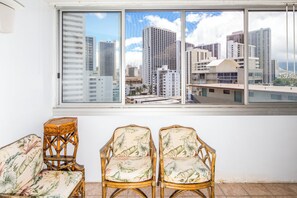 A room with a large window overlooking a cityscape, two wicker chairs with a tropical print, and a white wall. - Sundeck Studio w/ Kitchenette - Waikīkī Grand 1003 (Honolulu)
