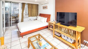 Hotellihuone, jossa on sänky, puinen jalusta, jonka päällä on televisio, lasinen sohvapöytä ja suuret ikkunat, joista avautuu näkymä kaupunkiin.