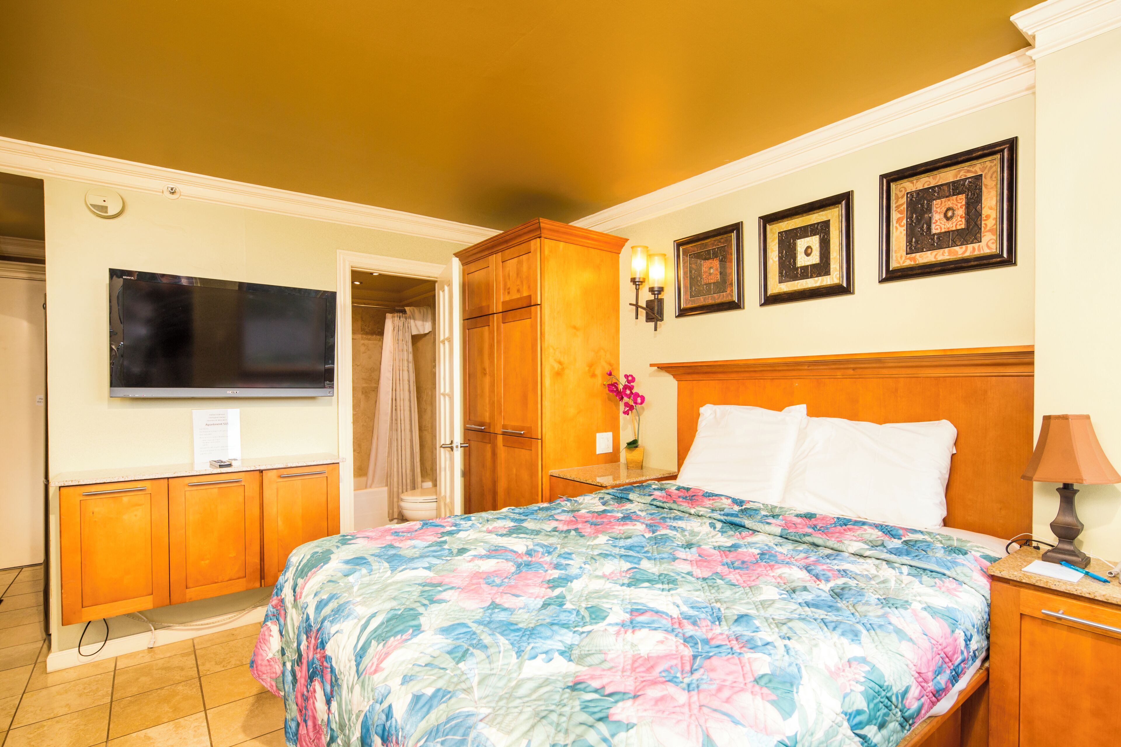 A hotel room with a bed, a television, wooden cabinets, and a wall-mounted lamp.