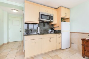 A kitchen with light wooden cabinets, a stainless steel microwave, a white refrigerator, a marble countertop, and a small wooden cabinet. - Ocean View Studio w/Kitchenette- Waikīkī Grand 709 (Honolulu)