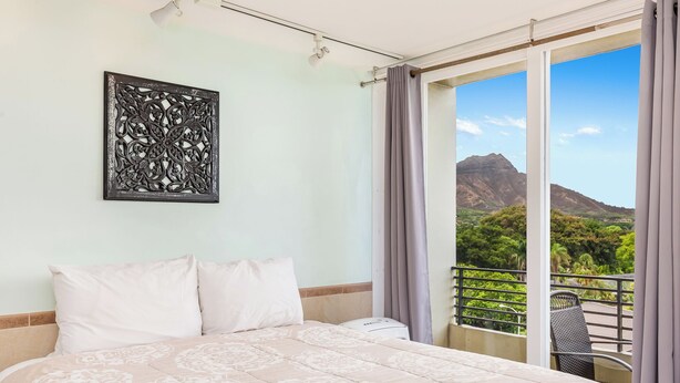 A hotel room with a bed, a wall-mounted artwork, a balcony with a view of mountains, and a chair. - Ocean View Studio w/Kitchenette- Waikīkī Grand 709 (Honolulu)