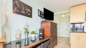 A dining area with a glass table, chairs, a TV mounted above, a wooden cabinet, and a kitchen area with cabinets and a sink. - Ocean View Studio w/Kitchenette- Waikīkī Grand 709 (Honolulu)