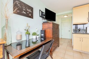 A dining area with a glass table, chairs, a TV mounted above, a wooden cabinet, and a kitchen area with cabinets and a sink. - Ocean View Studio w/Kitchenette- Waikīkī Grand 709 (Honolulu)