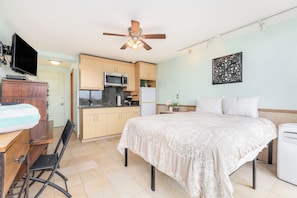 A bedroom with a bed, a desk, a chair, a TV, a ceiling fan, a microwave, a refrigerator, and a small kitchen area. - Ocean View Studio w/Kitchenette- Waikīkī Grand 709 (Honolulu)