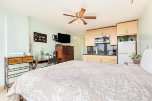 Een compacte kamer met een bed, bureau, kitchenette, plafondventilator en een televisie.