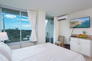 A hotel room with a large bed, a TV on the wall, a balcony with curtains, and a fruit basket on the counter. - Ocean View Studio with Kitchenette - Waikīkī Grand #910 (Honolulu)