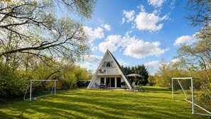Ein dreieckiges Haus mit Terrasse und Fußballtor auf einer grünen Wiese.