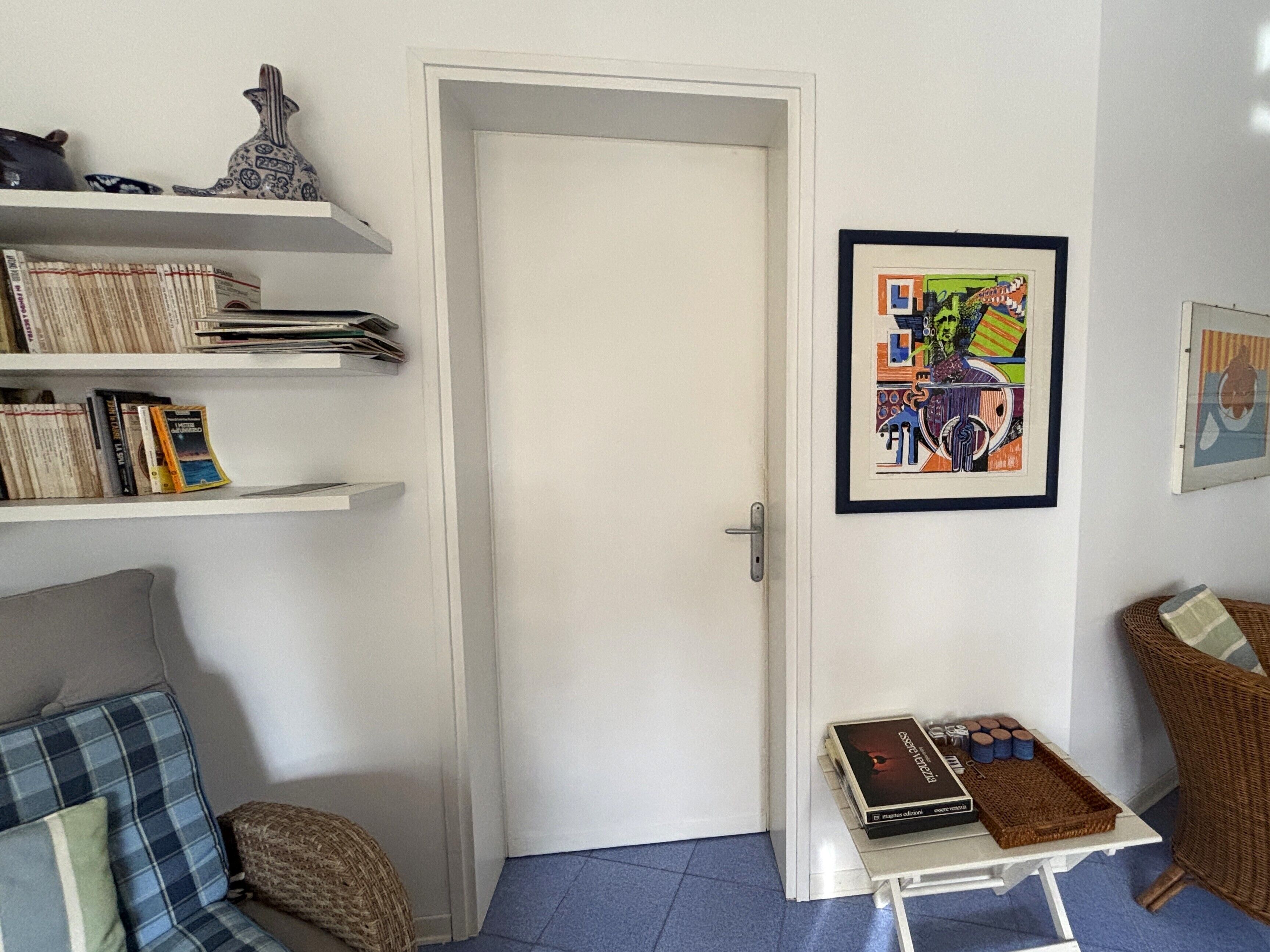 Una habitación con una puerta blanca, una estantería con libros y objetos decorativos, un cuadro abstracto enmarcado, un sillón de mimbre y una pequeña mesa con un libro y velas.