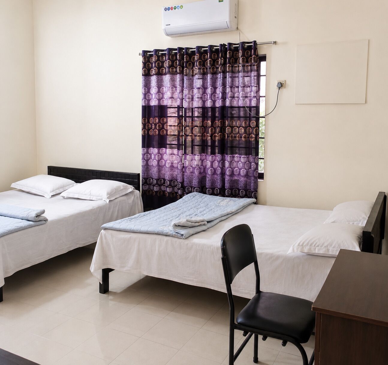 A hotel room with two single beds, a desk, a chair, an air conditioner, and a patterned curtain.