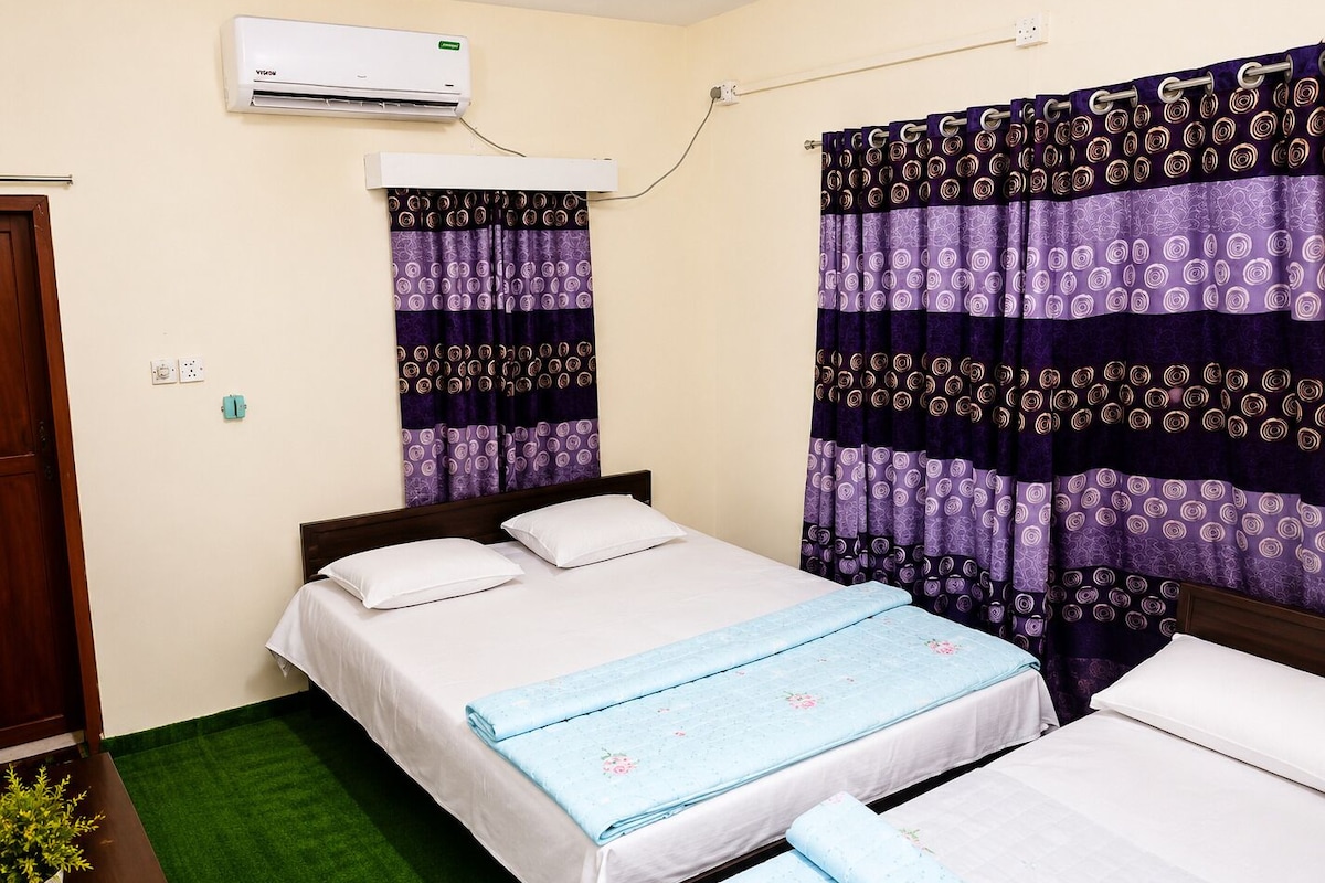 Un dormitorio con dos camas, alfombra verde, aire acondicionado y cortinas moradas.