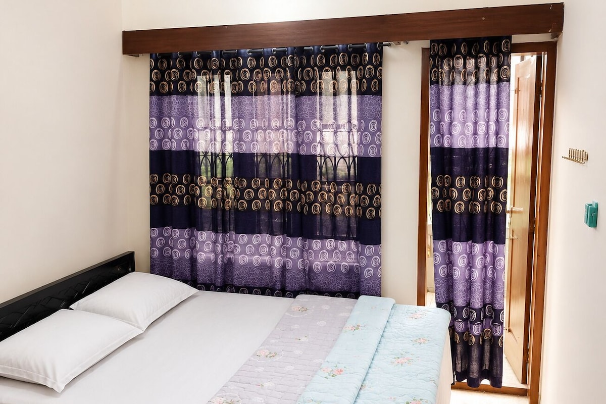 Una cama con ropa blanca, un colcha floral azul y cortinas moradas estampadas.