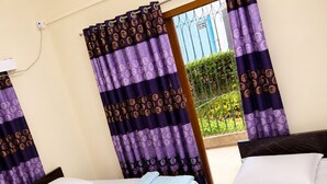 Habitación de hotel con dos camas, una ventana con cortinas moradas y vistas a zonas verdes.