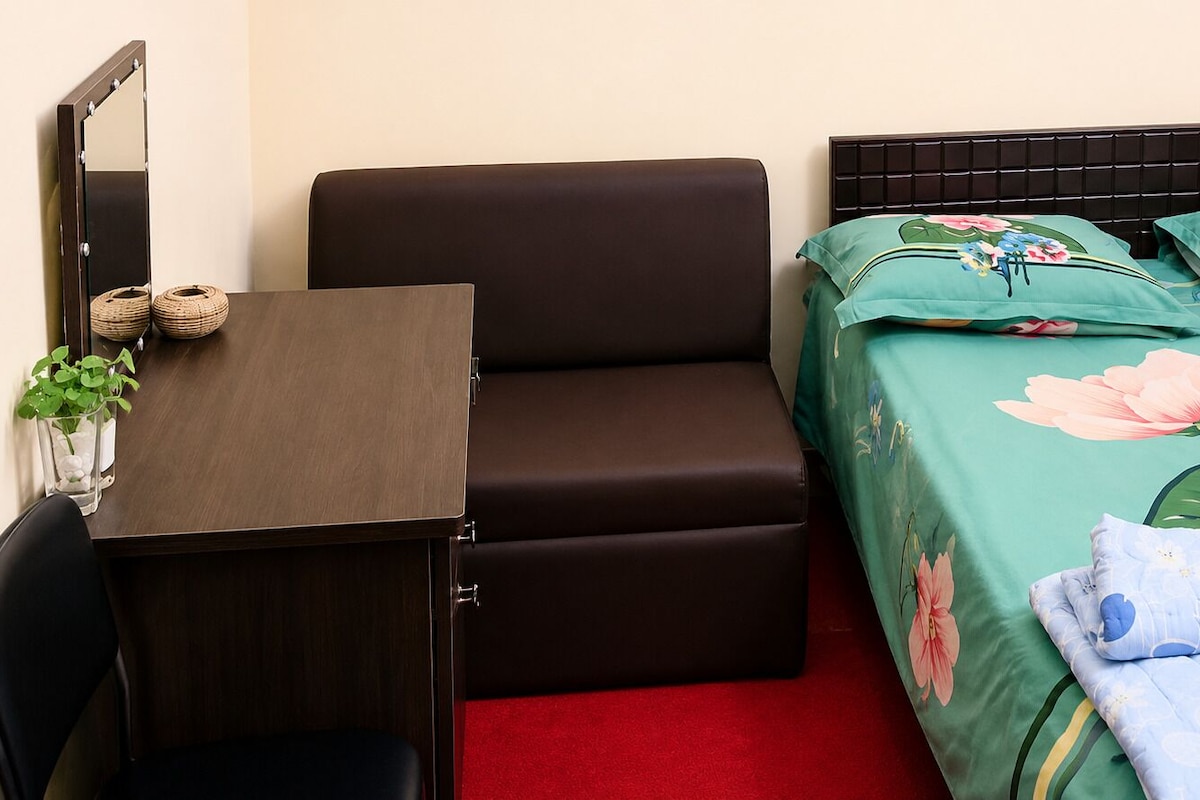 Un dormitorio con cama, escritorio, silla, espejo y una planta pequeña.