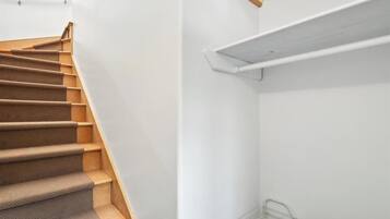 A staircase with carpeted steps and a metal handrail, leading to a white wall with a wooden shelf and a metal wire shelf.