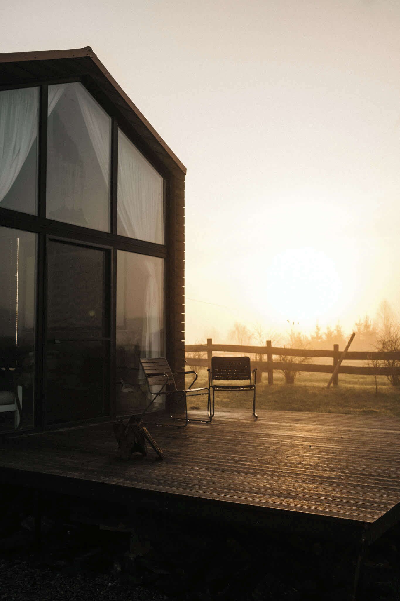 Una terraza de madera con una silla y un banco, un edificio de cristal con cortinas y una valla al fondo.