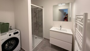 Une salle de bain moderne dotée d’une douche à parois de verre, d’un lave-linge encastré, d’un meuble-lavabo blanc avec un miroir et d’un porte-serviettes.