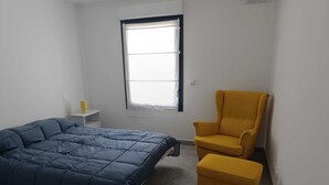 Une chambre à coucher avec un lit, un fauteuil jaune, une petite table avec une lampe et une fenêtre.