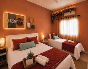 A hotel room with two beds, a wall clock, a decorative wall piece, a window with curtains, and a small table with a plant and a tray of towels.
