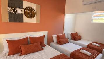 Ein Hotelzimmer mit vier Betten, jedes mit einem weißen Kopfteil und orangener Bettwäsche. Darüber hängen Bilder an der Wand, und an der Wand ist eine Klimaanlage angebracht.