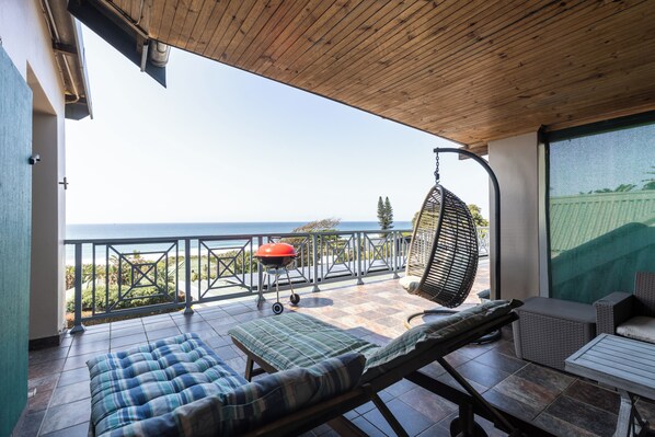 A balcony with a view of the sea, a hanging chair, a grill, and lounge chairs.
