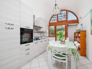 A modern kitchen with white cabinetry, a built-in oven, and a dining area with a table set for four. - De Vivo Realty - Golden Garden Sorrento. (Meta)