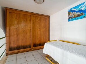 A bedroom with a wooden wardrobe, a bed with a white mattress, and a wall-mounted painting of a coastal scene. - De Vivo Realty - Golden Garden Sorrento. (Meta)