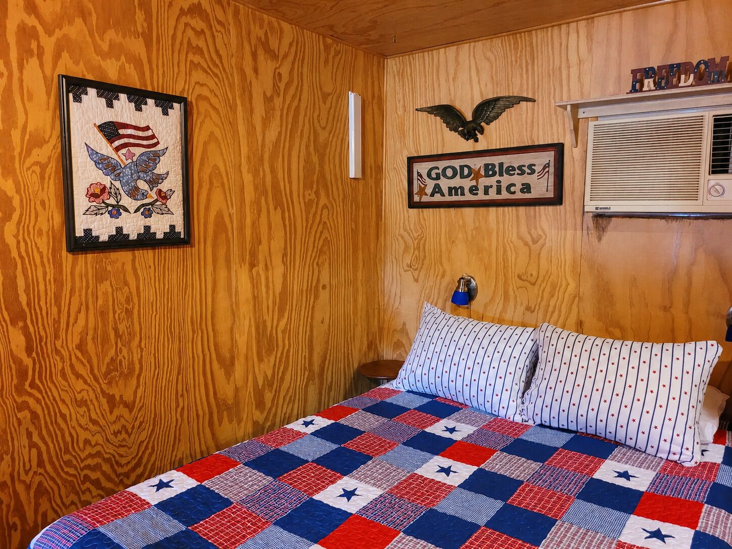 Ein Zimmer mit Holzvertäfelung, einem Bett mit einer quiltartigen Steppdecke mit Sternenmuster, einer gerahmten Kunstgrafik der amerikanischen Flagge und einem Wandschild mit der Aufschrift 'GOD Bless America'.