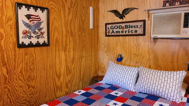 Una habitació amb panells de fusta, un llit amb col·cha amb dibuixos de estrelles, una obra d'art amb una bandera americana emmarcada, i un cartell a la paret que diu 'GOD Bless America'.