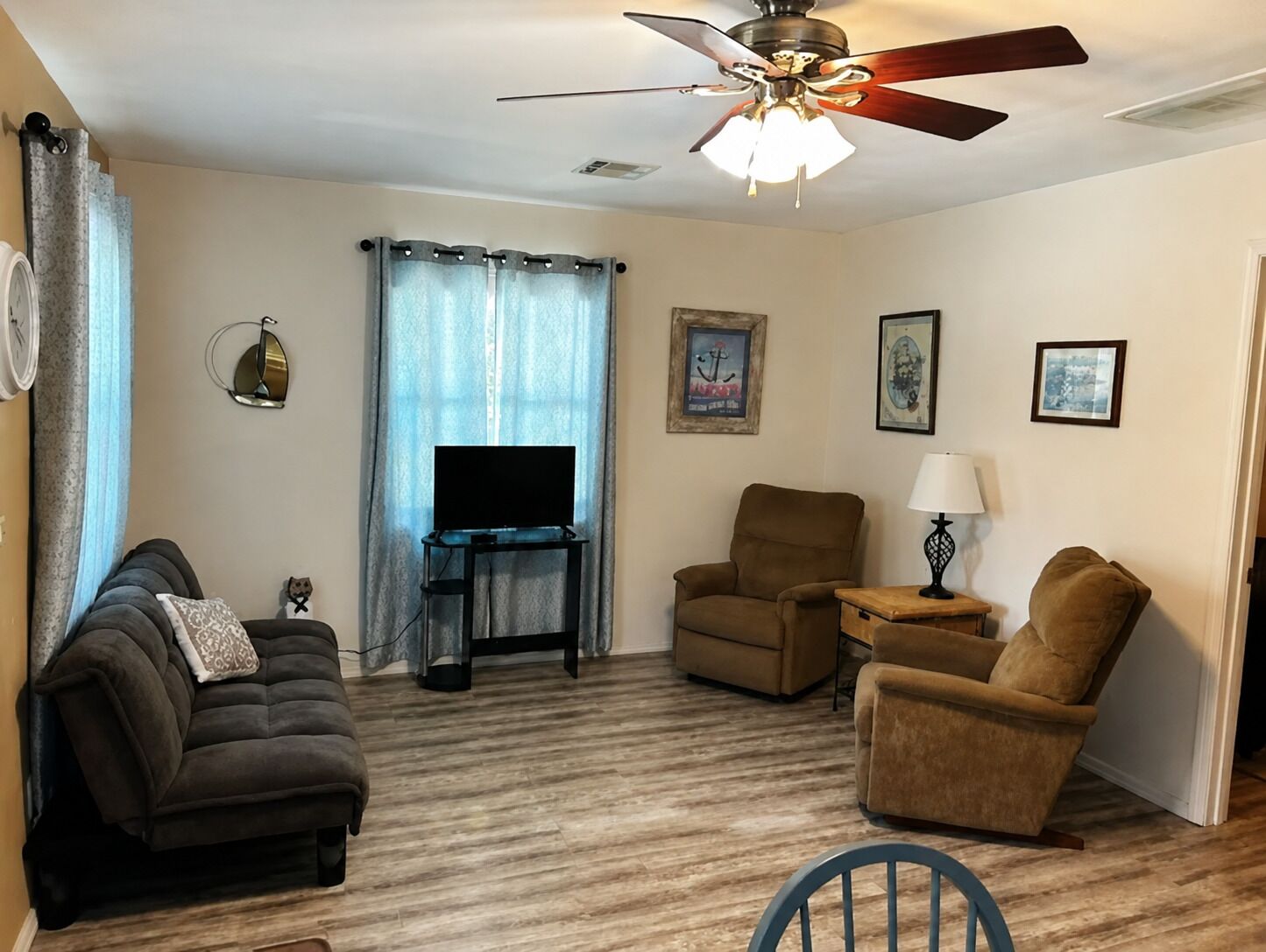 A living room with a ceiling fan, a television on a stand, two armchairs, a sofa, and a lamp on a side table.